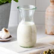 Creamy white caesar dressing in a small glass jar with capers in the front and a garlic head at the back.
