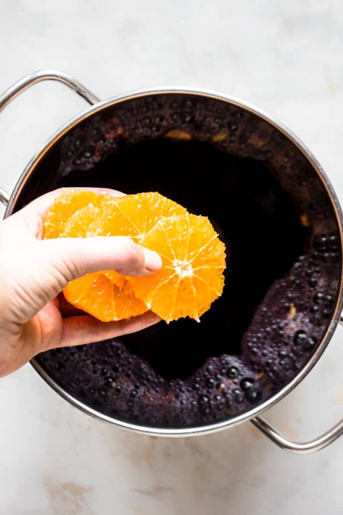 Hand adding orange slices to the pot with grape juice.