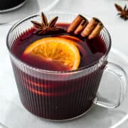 Ribbed glass cup filled with hot wine, orange slices and two cinnamon sticks.