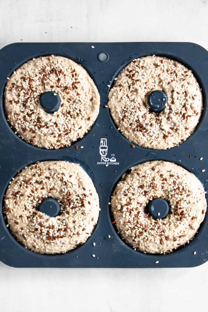 Bagels topped with seeds in a dark blue doughnut pan before baking.