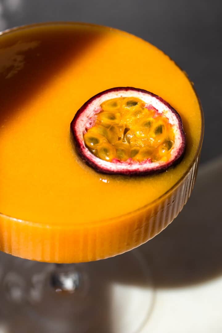 Close-up photo of half a passion fruit on a vibrant orange cocktail in a coupe.
