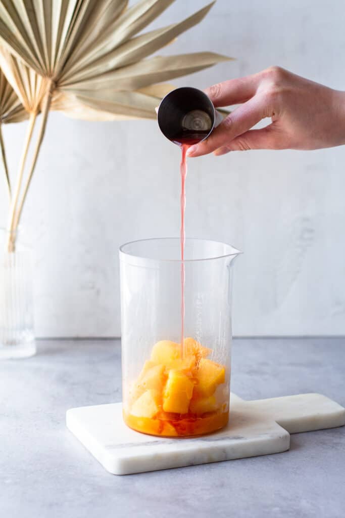 Hand pouring passion fruit liqueur out of a jigger into a container with frozen mango.