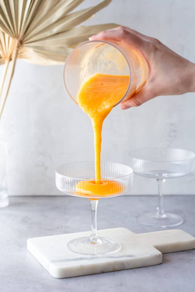 Hand pouring frozen orange-colored cocktail into a coupe glass.