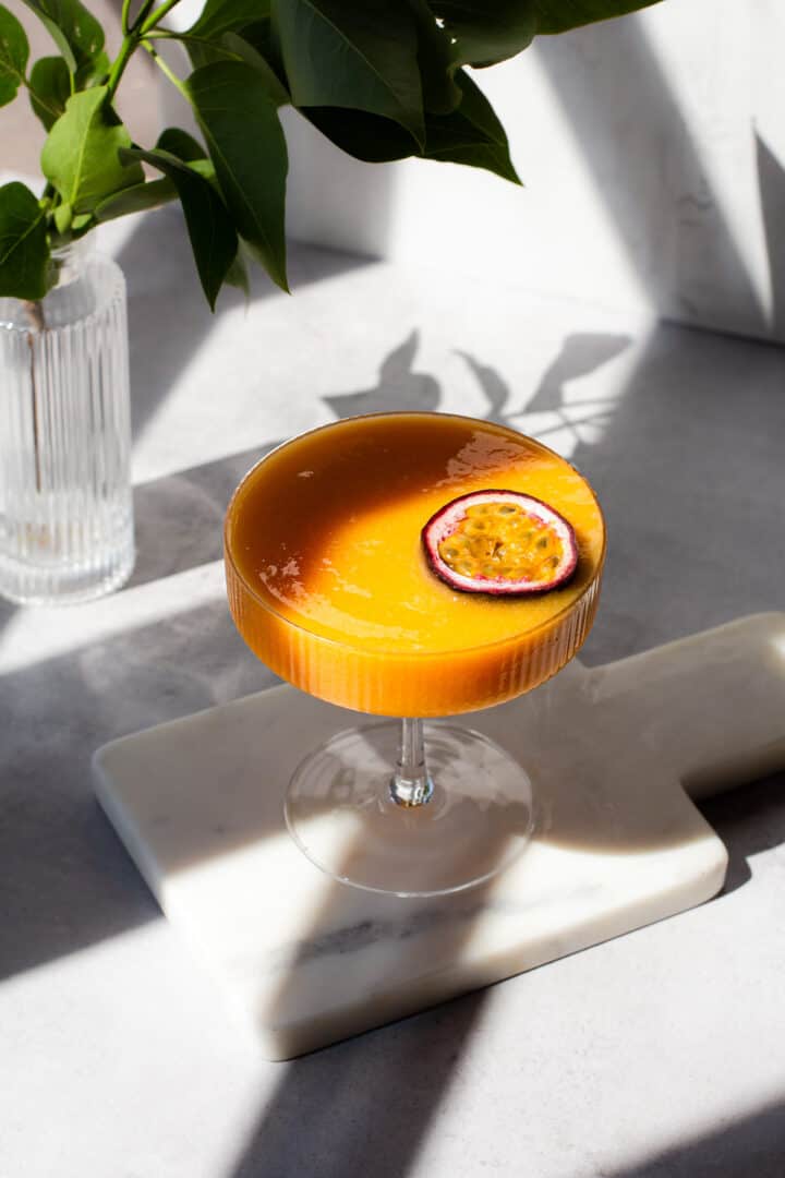 Coupe glass filled with orange cocktail and garnished with half a passion fruit on a on a marble board.