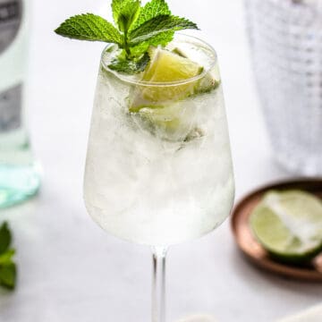 Stemmed wine glass filled with a translucent cocktail, garnished with crushed ice, lime and mint.