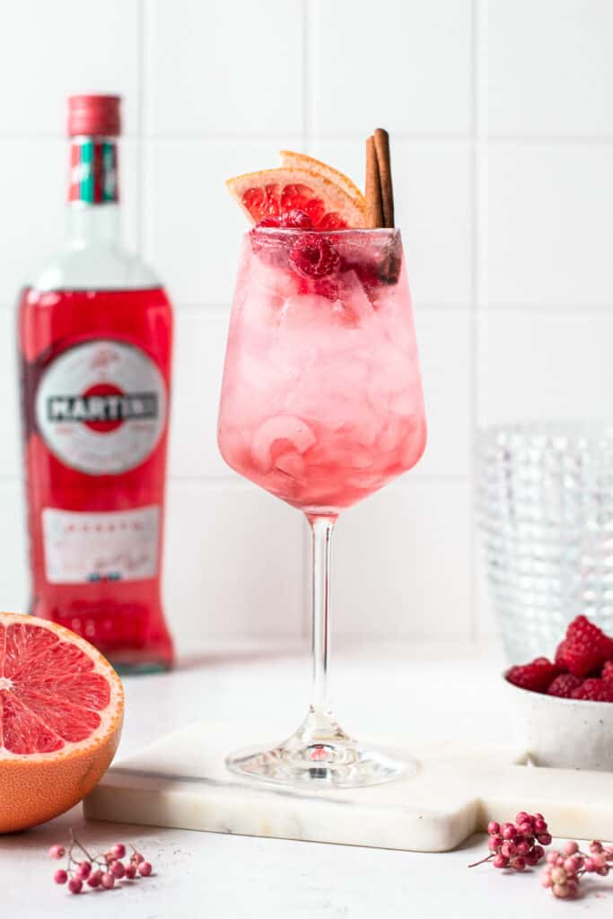 Pink cocktail garnished with grapefruit, raspberries and a cinnamon stick.