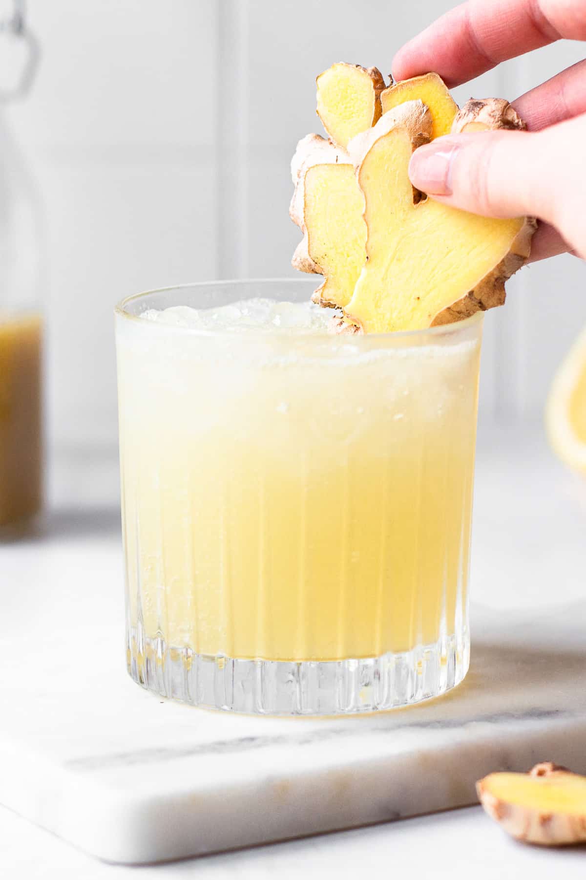 Hand adding to slices of fresh ginger to a low-rise glass.