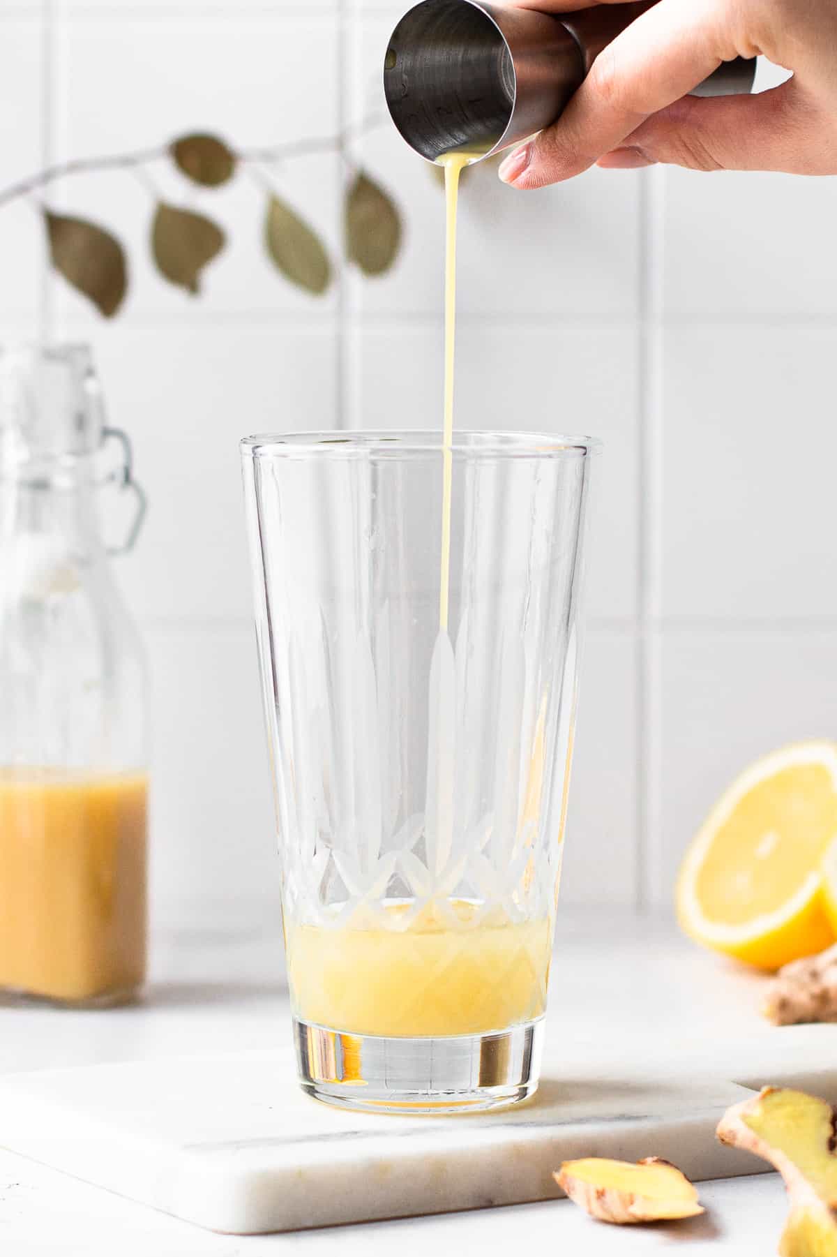 Hand pouring ginger syrup into a glass cocktail shaker.