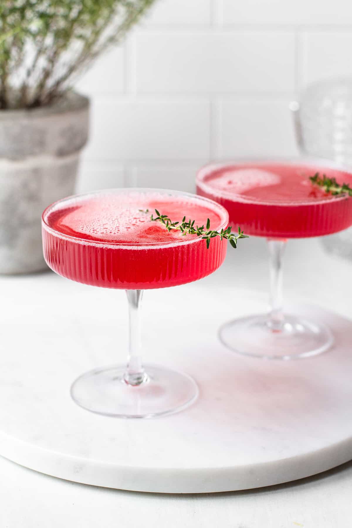Pink pomegranate elderflower cocktail in a ribbed coupe glass on a round marble board.