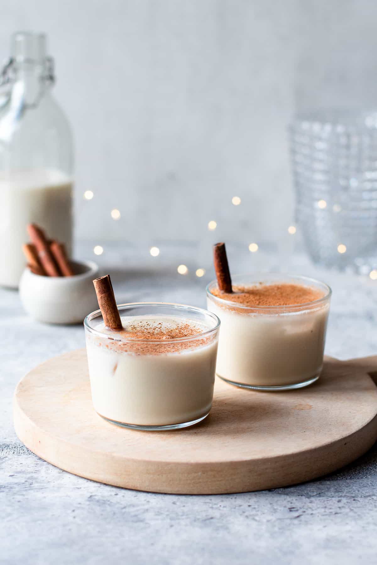 Two coconut milk cocktails with a cinnamon stick in it on a wooden board. Christmas lights in the background.