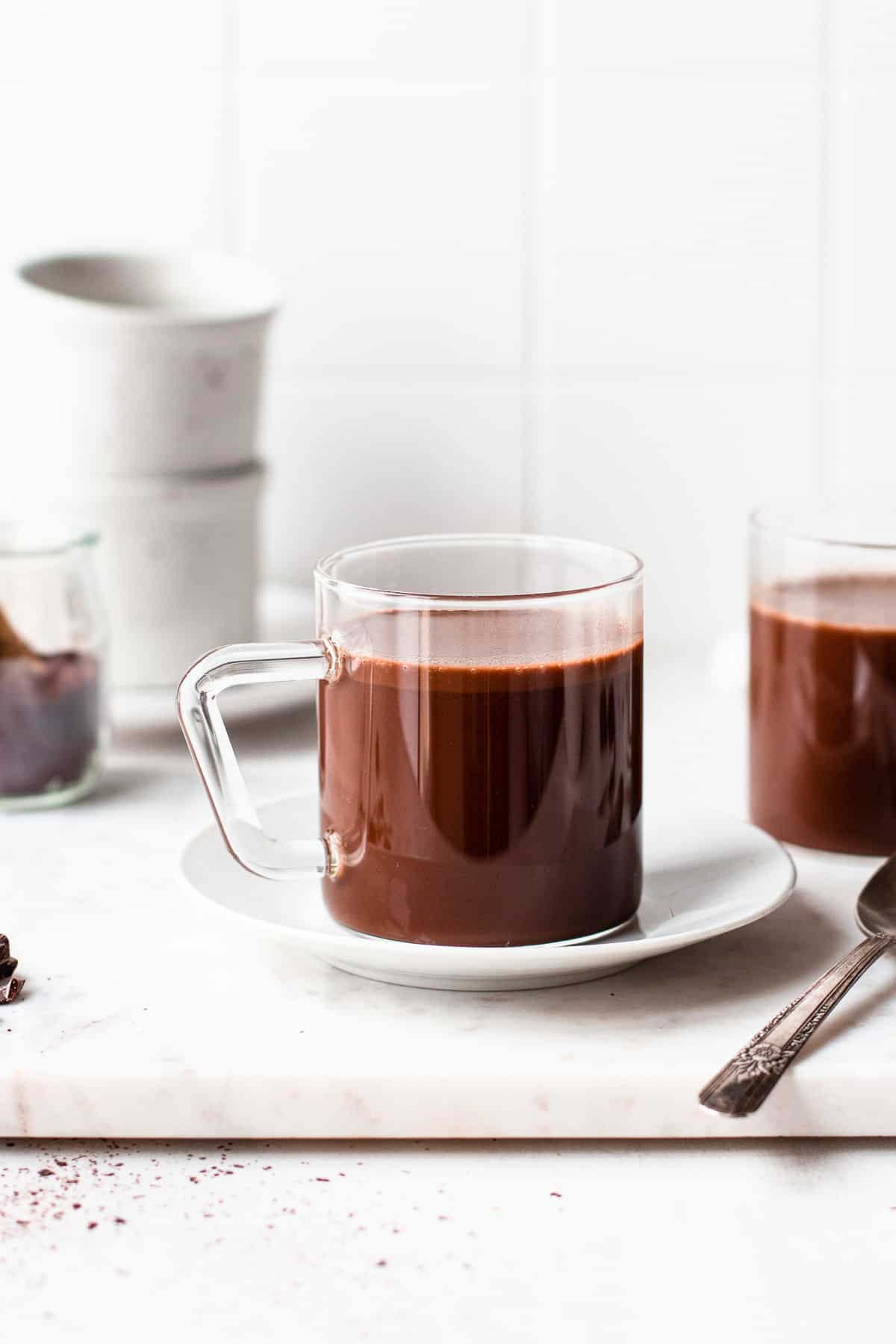 Glass mug filled with hot chocolate in front on a white tile background.