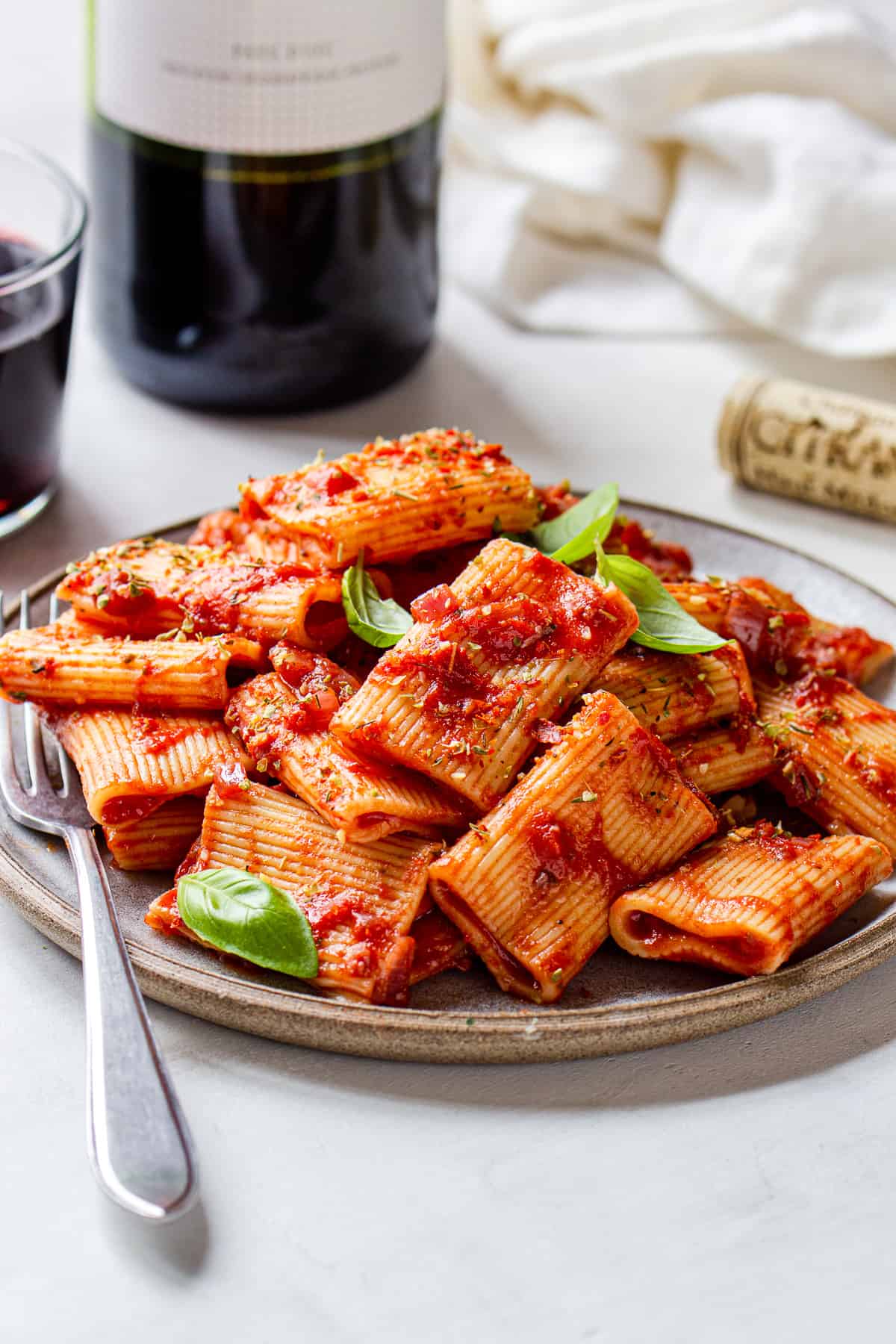 Plate with rigatoni pasta covered in red sauce and wine in the background.