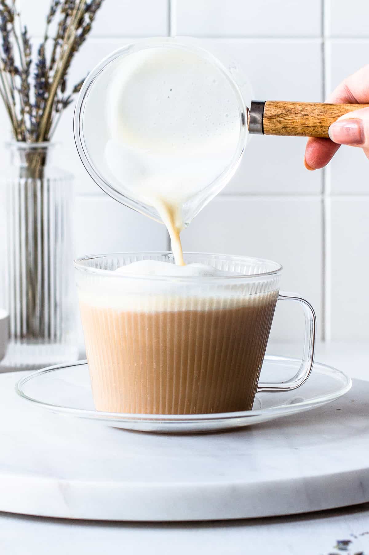 Hand pouring frothed milk into a ribbed glass cup with coffee.