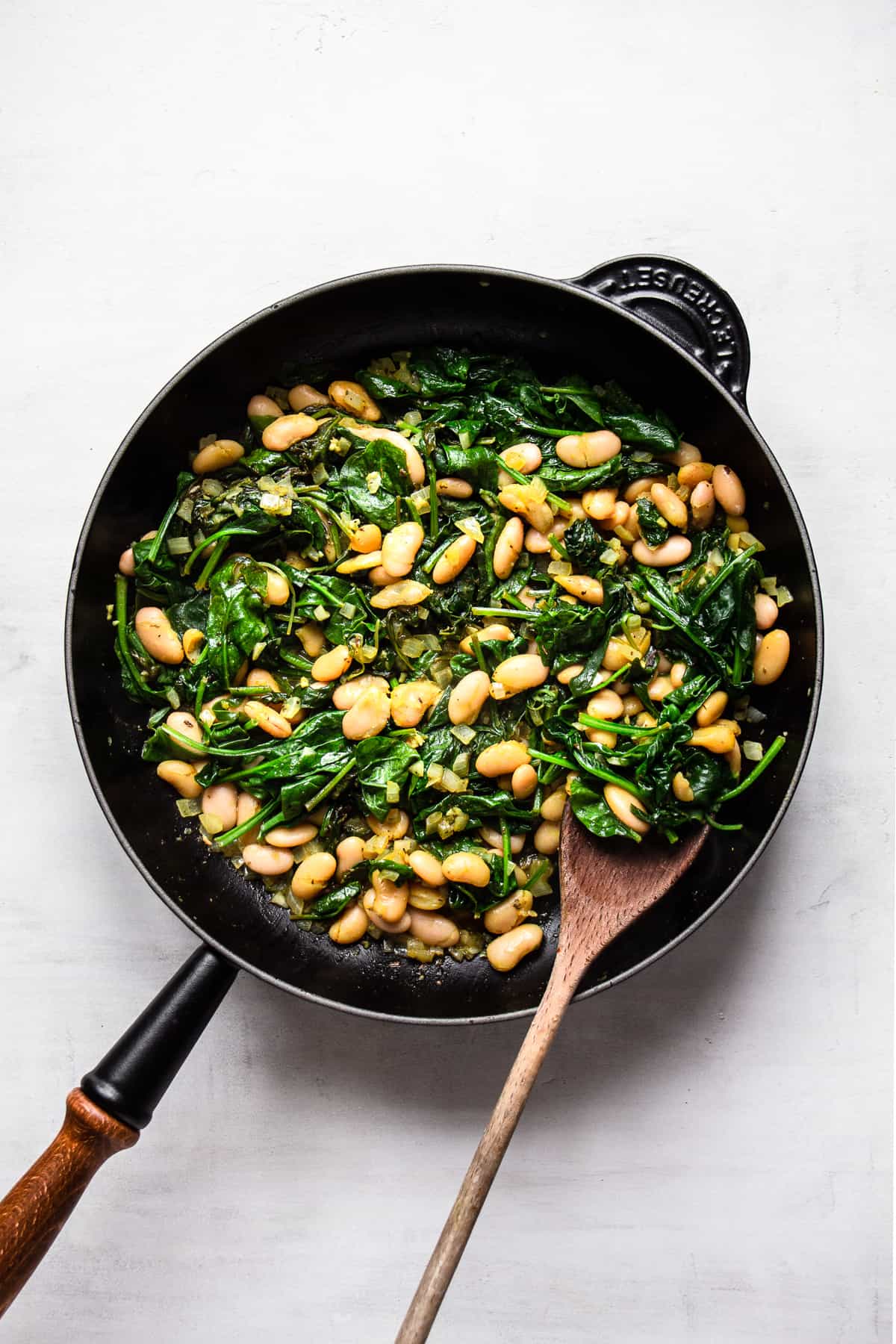 White beans and cooked spinach in a black pan with wooden handle.