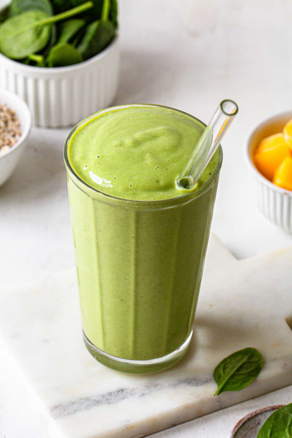 Green spinach smoothie in a glass and a bowl with frozen mango in the background.