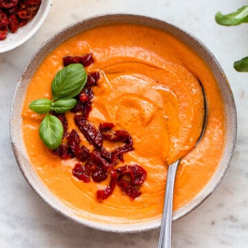 Grey ceramic bowl with bright orange tomato soup topped with sun-dried tomatoes and fresh basil.