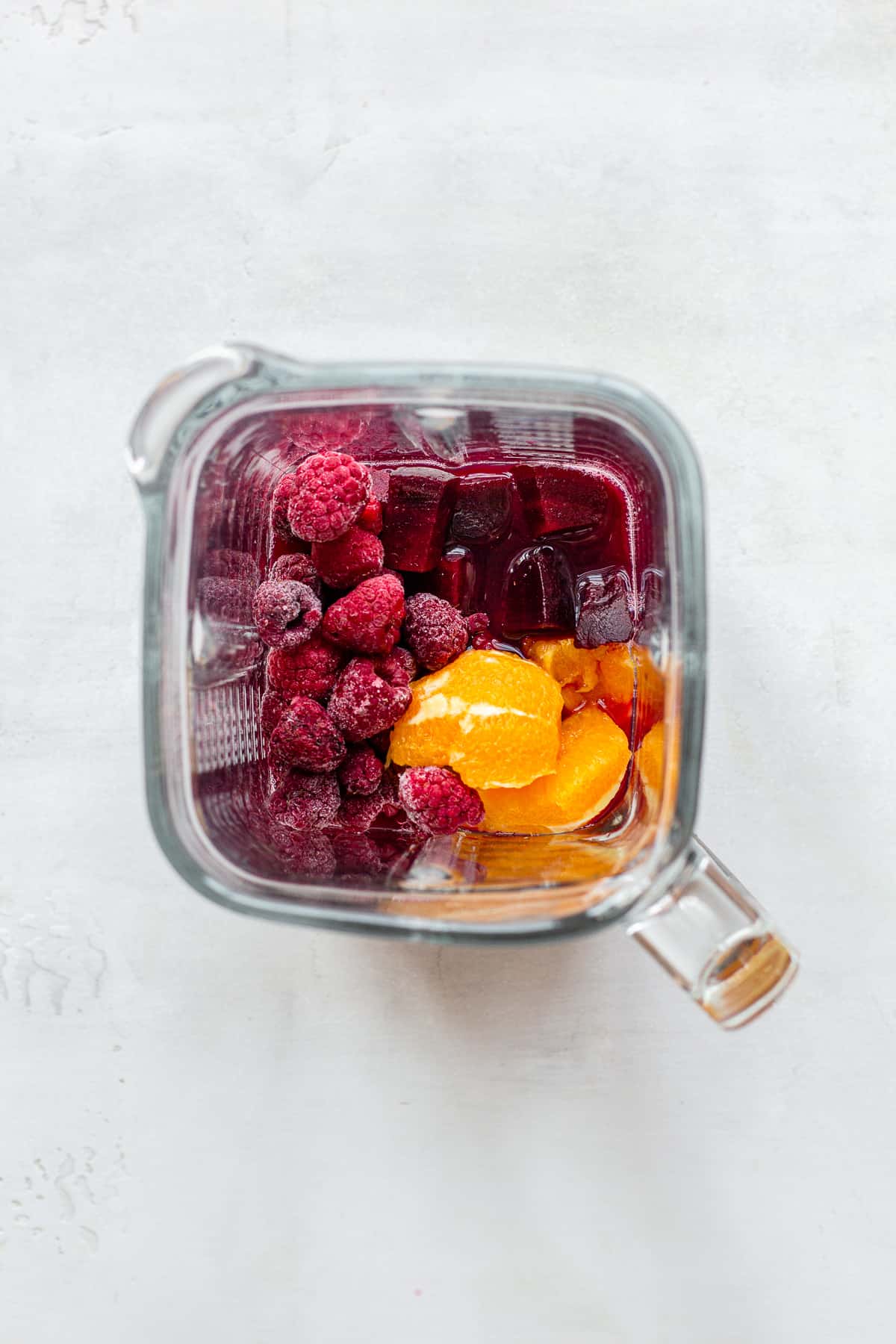 Blender jug with orange quarters, frozen raspberries, beetroot and cranberry juice.