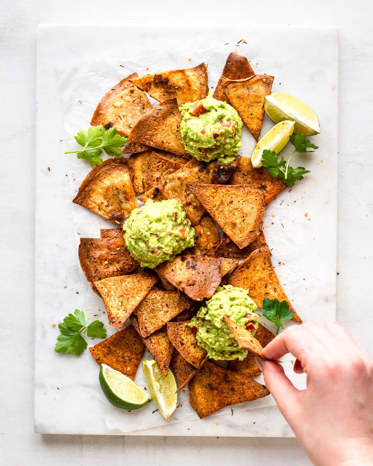 Hand dipping a nacho in some guacamole on a white marble platter.