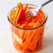 Fork taking ginger thin pickled carrot sticks out of a glass jar.