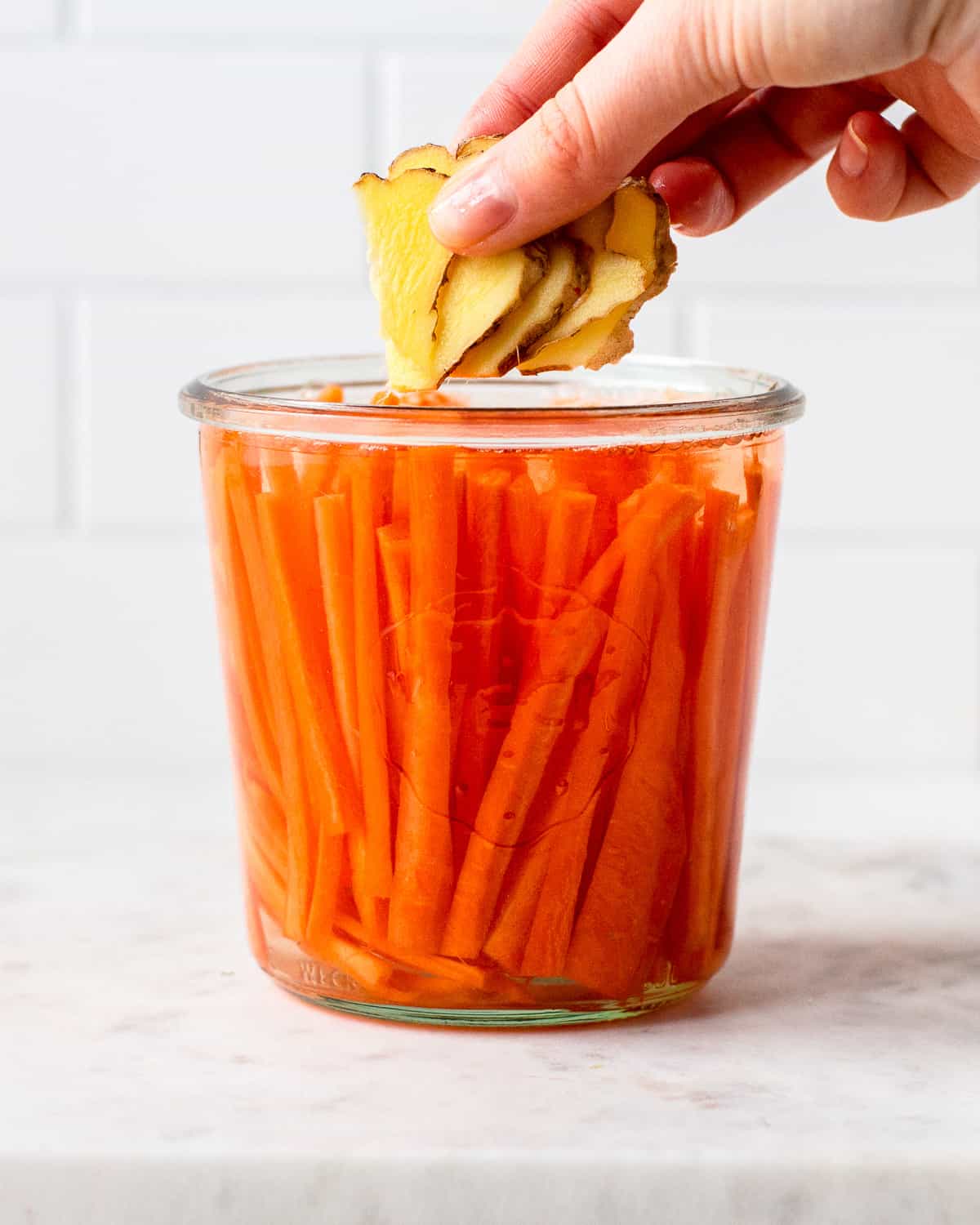Hand adding sliced ginger to a glass jar with carrot sticks.