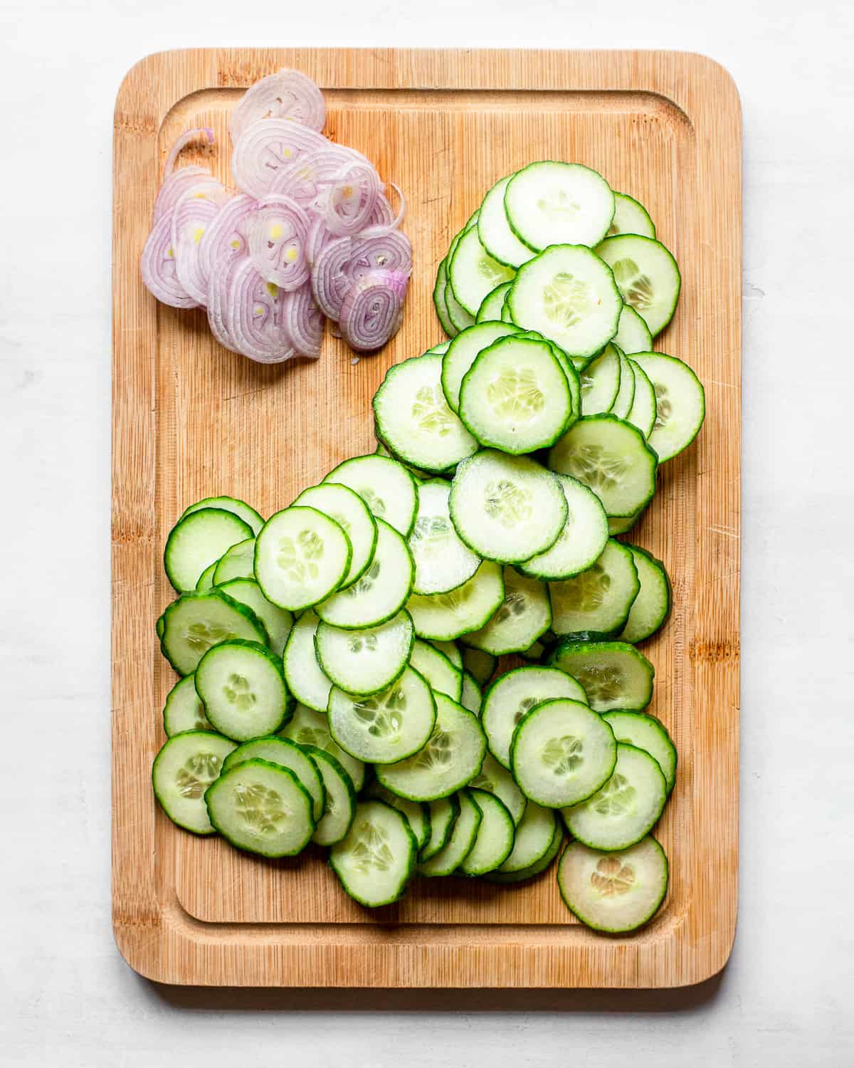 Sliced cucumbers and sliced shallots on a wooden cutting board.