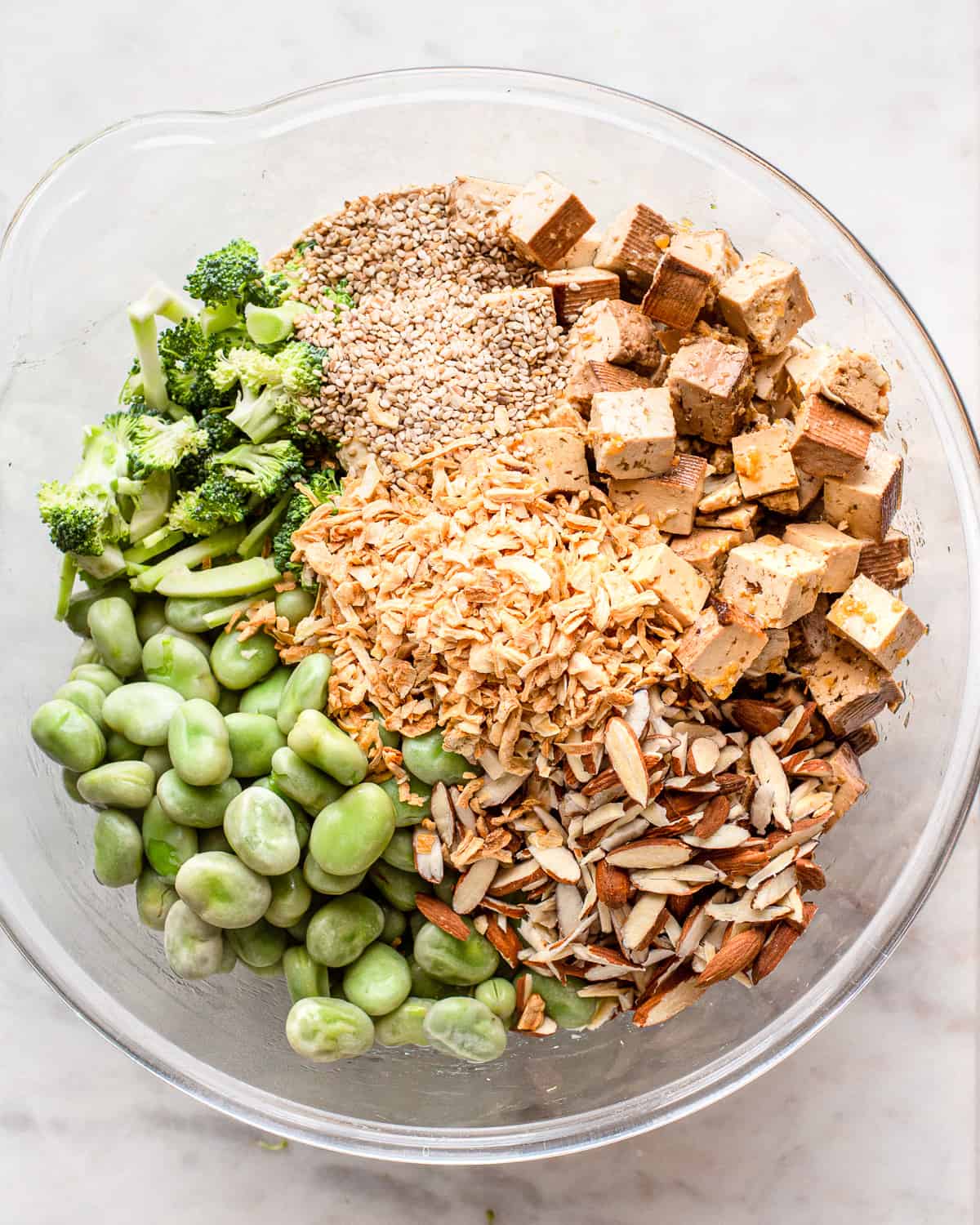 Ingredients for a broccoli, tofu, and edamame bean salad in a large glass bowl.