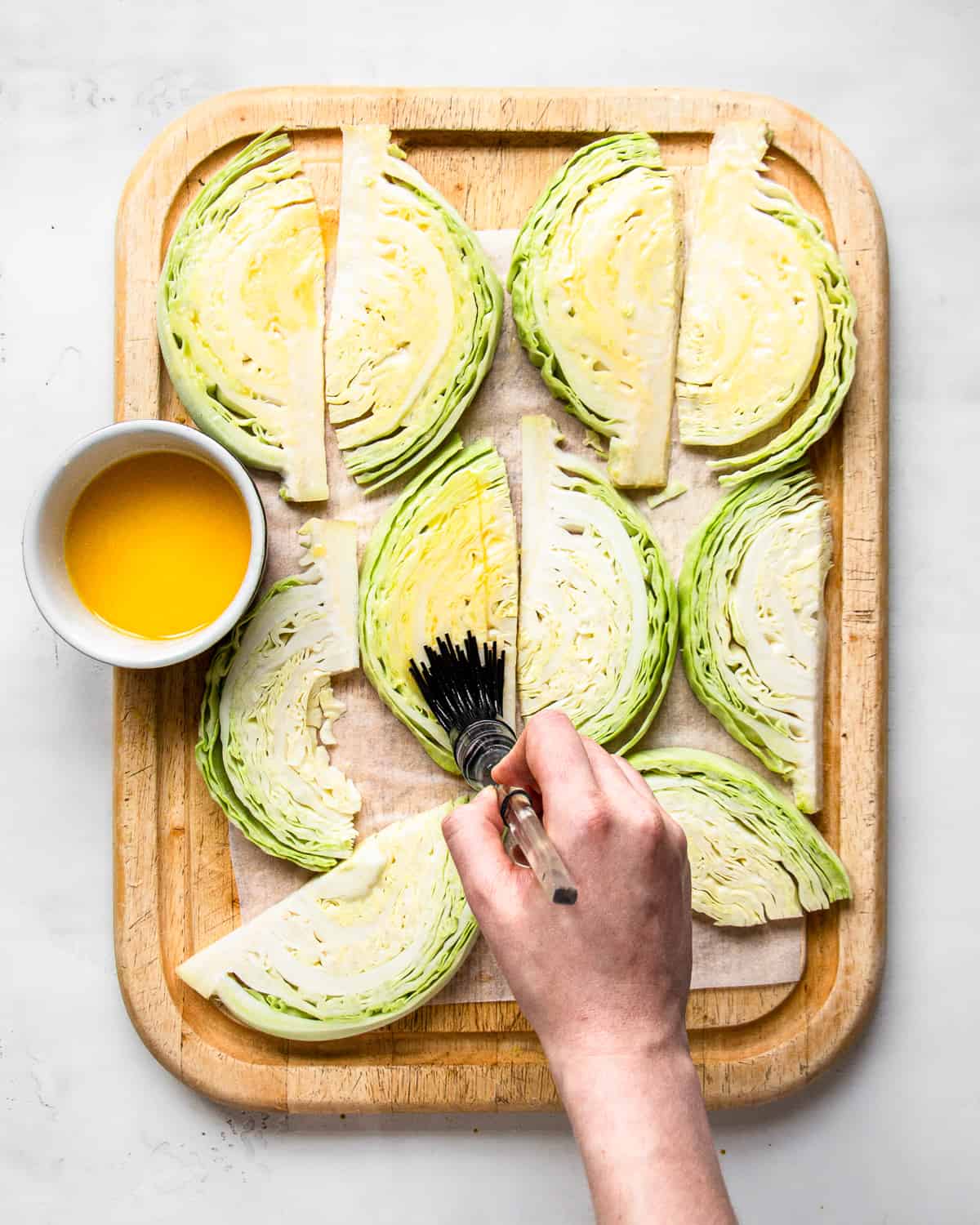 Hand brushing melted butter on cabbage wedges on a wooden cutting board.