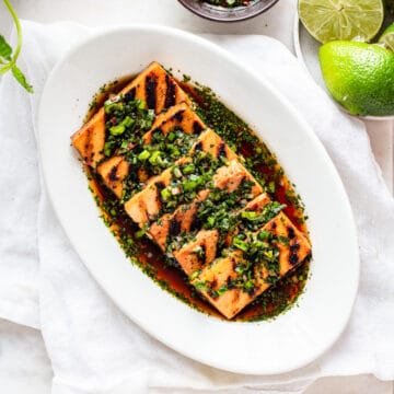Tofu planks with grill marks on a white oval dish covered with a brown sauce and green herbs.