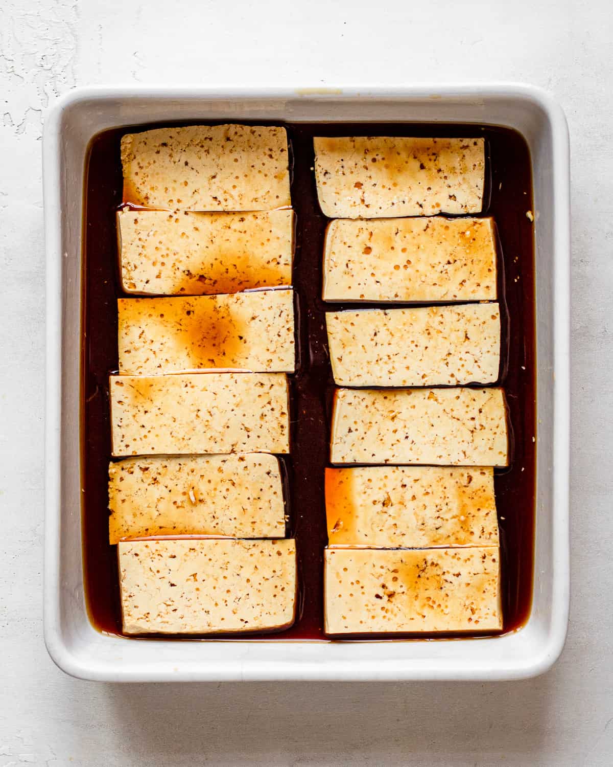 12 tofu planks in a soy sauce mixture in a white rectangular baking dish.