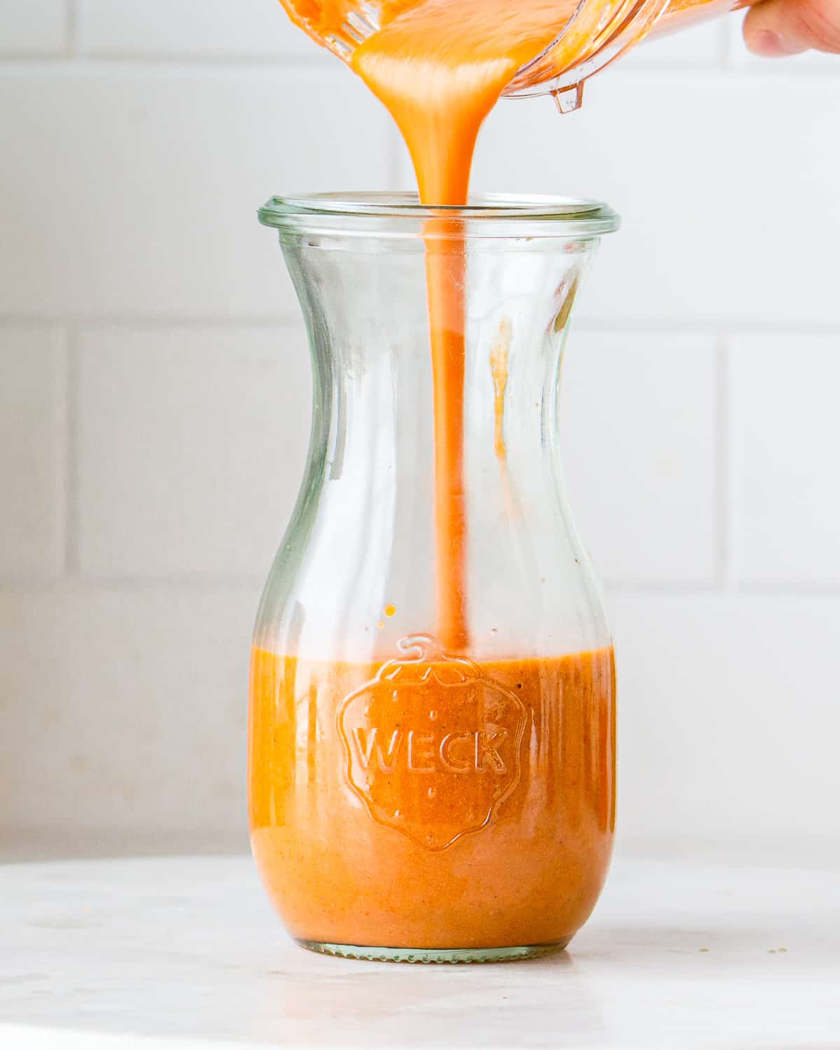 Hand pouring a creamy red-orange dressing into a small glass jar.