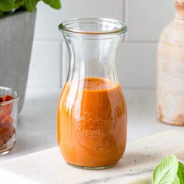 Red-orange salad dressing in a small glass jar with basil in the front and sun-dried tomatoes in the back.