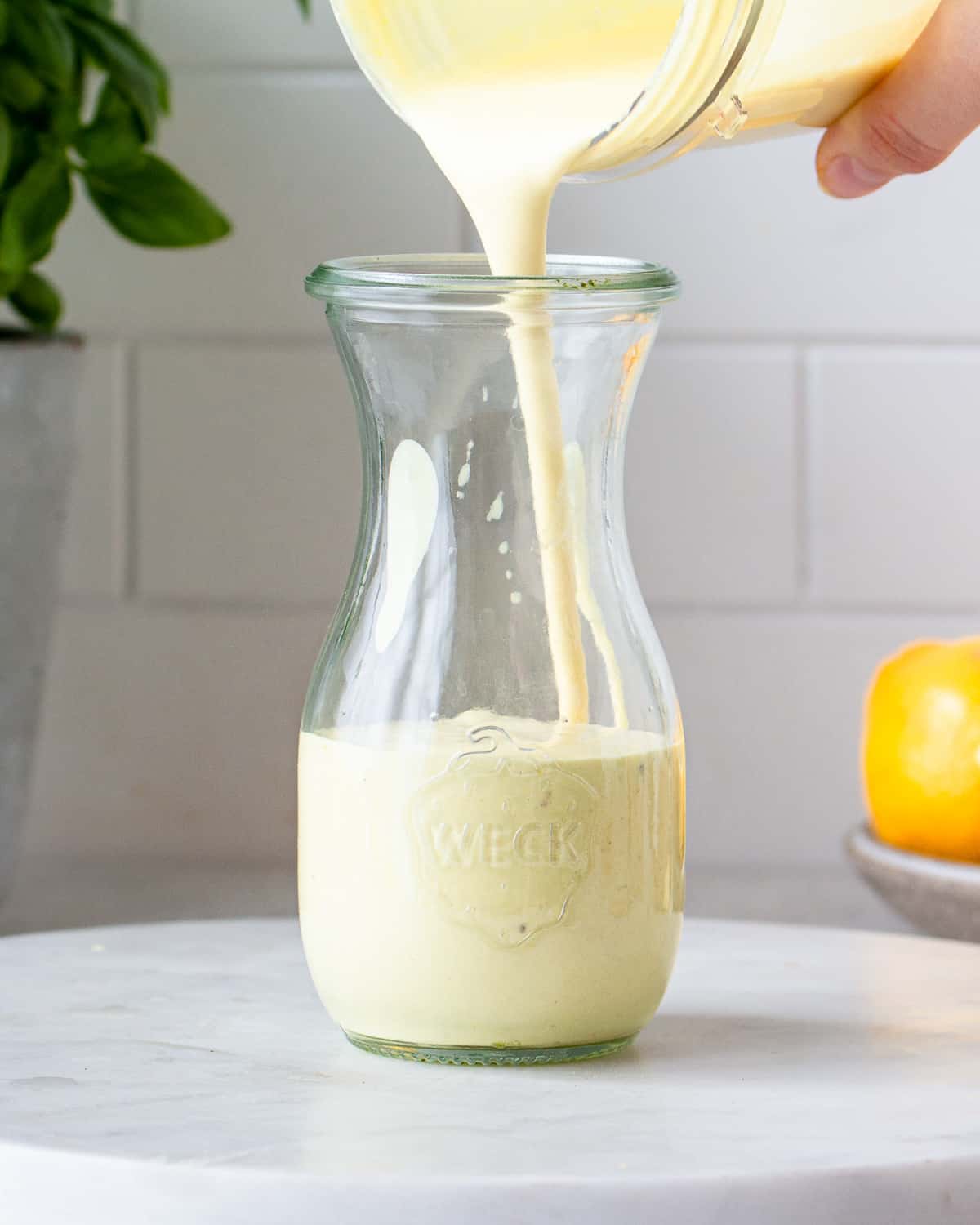 Hand pouring a creamy, light yellow salad dressing into a small glass jar.
