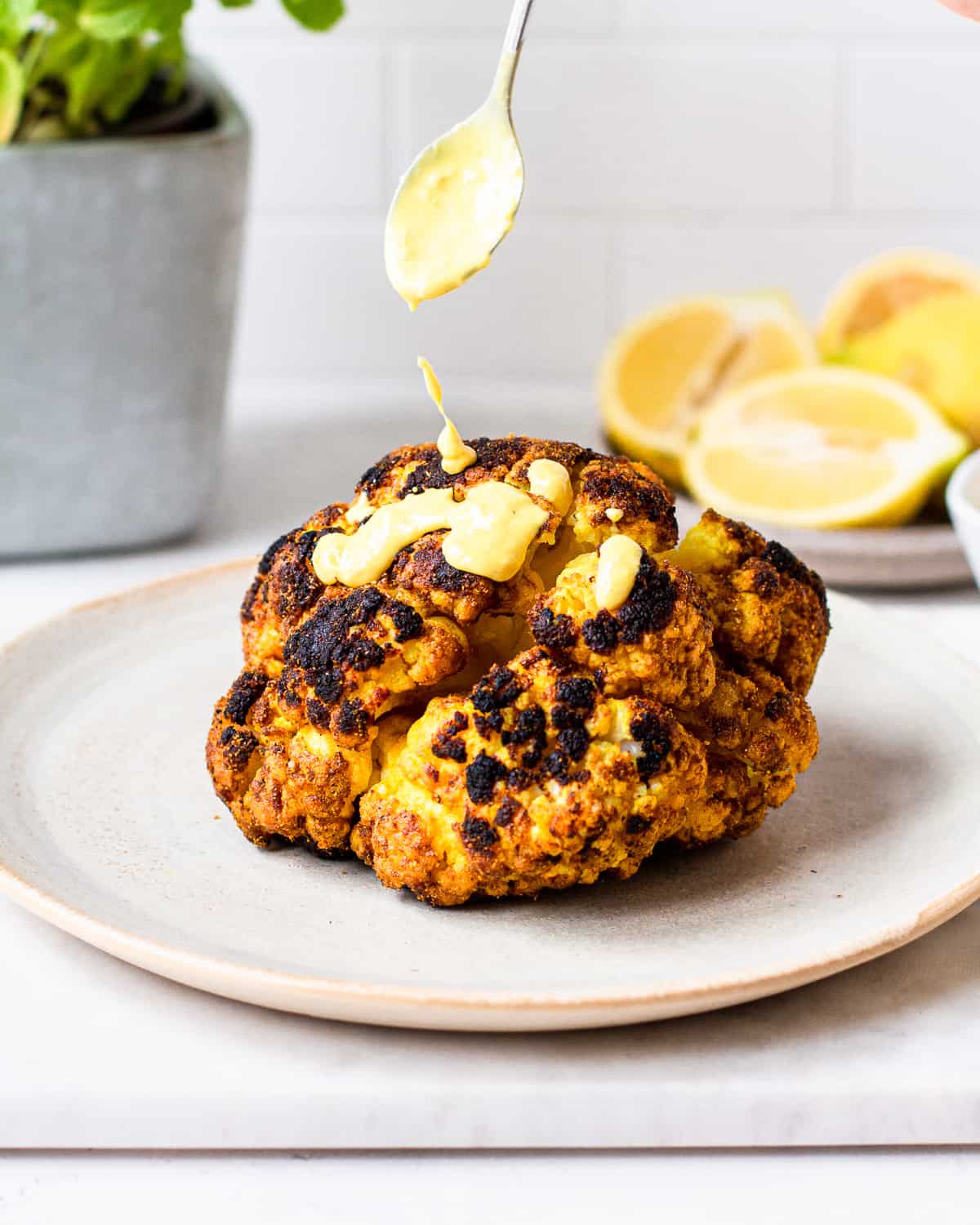 Spoon drizzling a yellow tahini sauce onto a grilled cauliflower head.