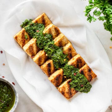Six pieces of golden brown tofu arrange in a row on an oval white plate with chimichurri on top.