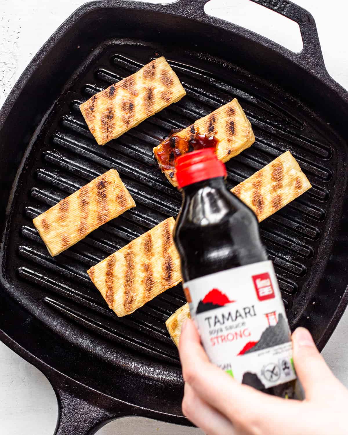 Hand pouring tamari over tofu pieces in a cast iron grill pan.