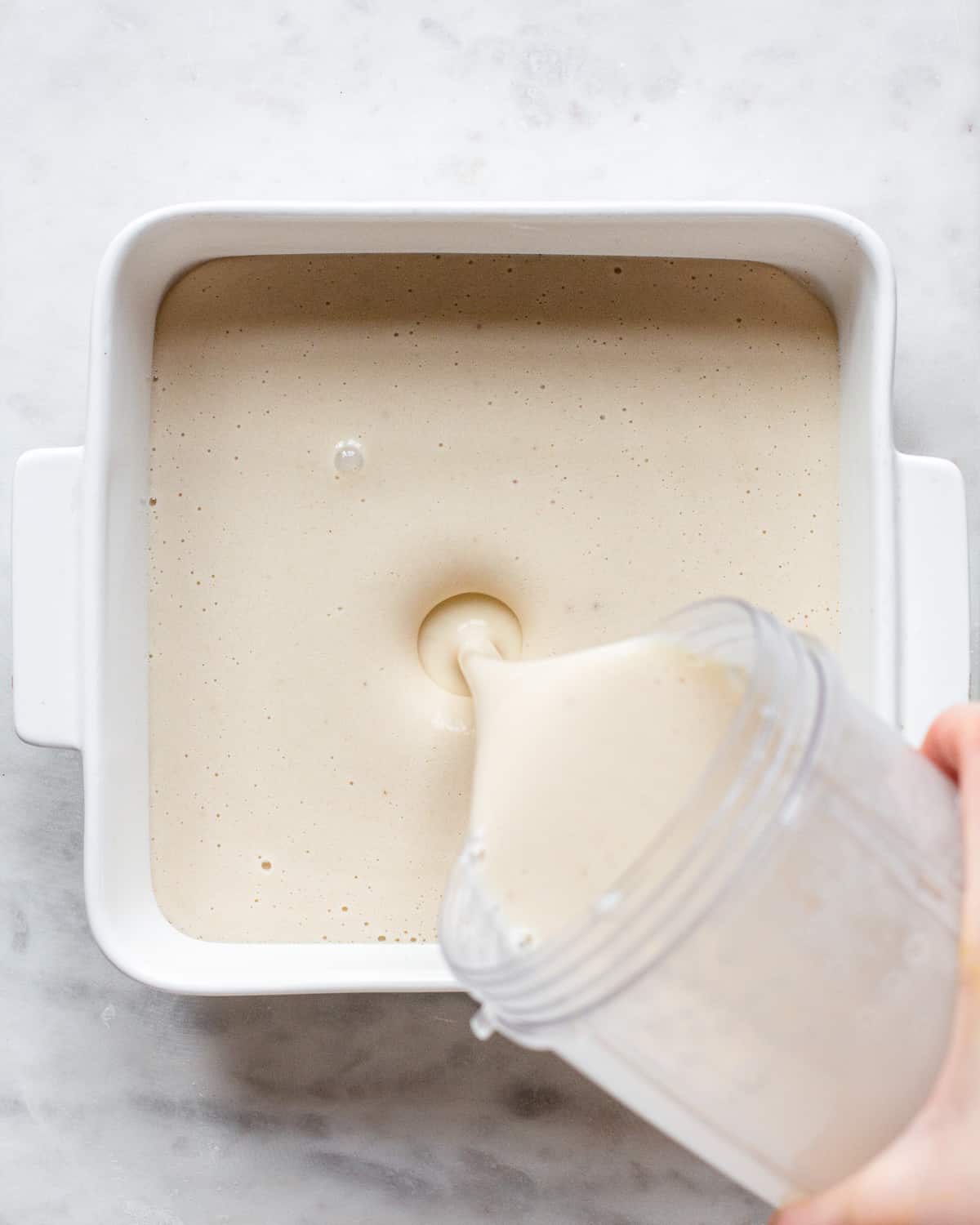Hand pouring blended banana mixture into a white baking dish.