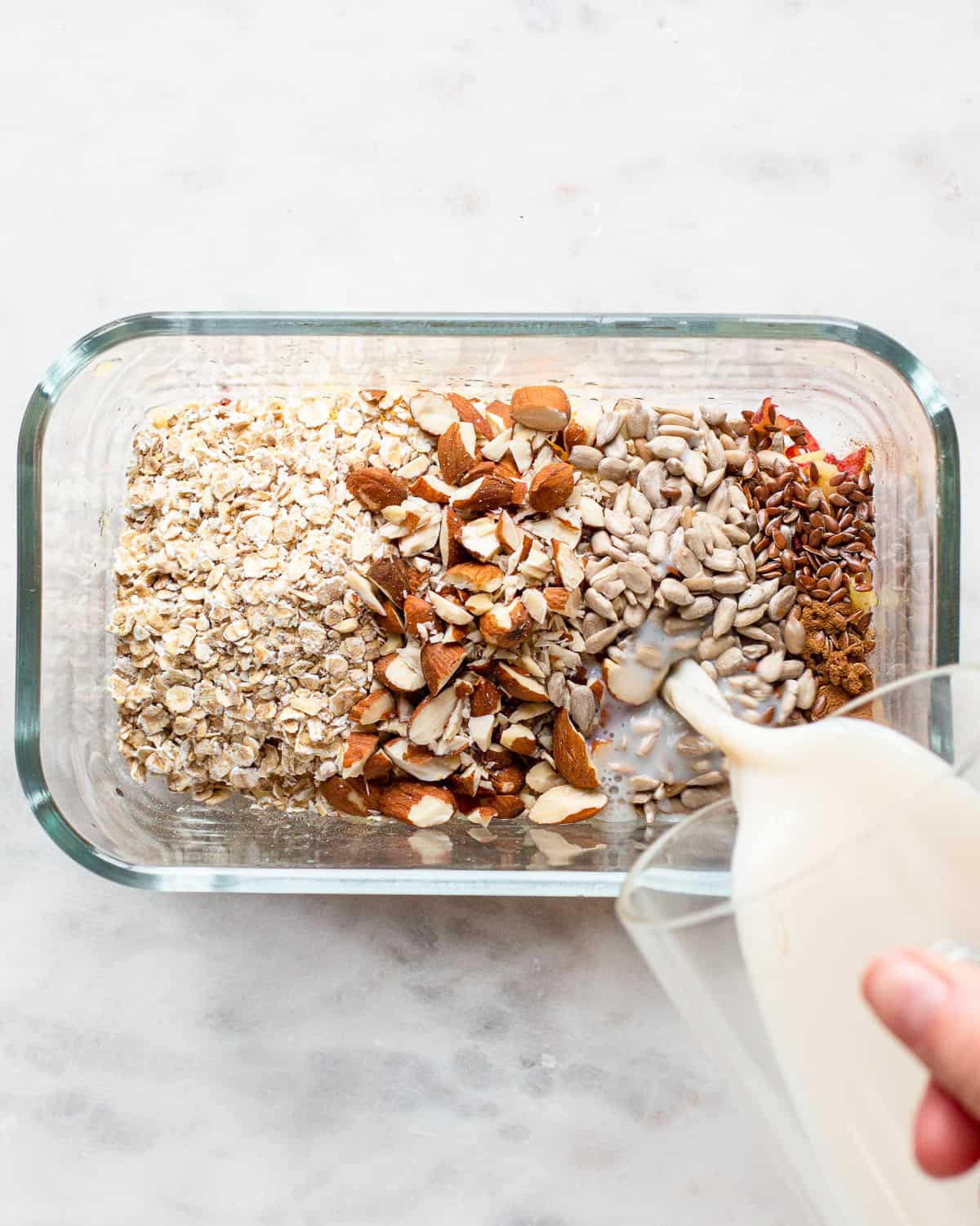 Hand pouring milk into a glass container with oats, nuts, ands seeds.