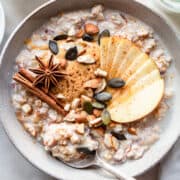 Grey ceramic bowl with bircher muesli topped with sliced apple, nut butter, a cinnamon stick, and a star anise.