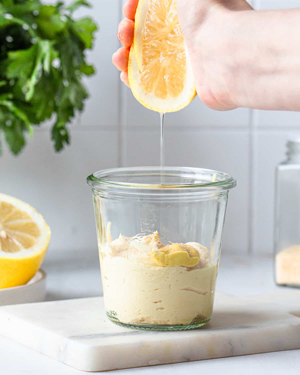 Hand adding lemon juice to a glass jar with hummus and mustard.