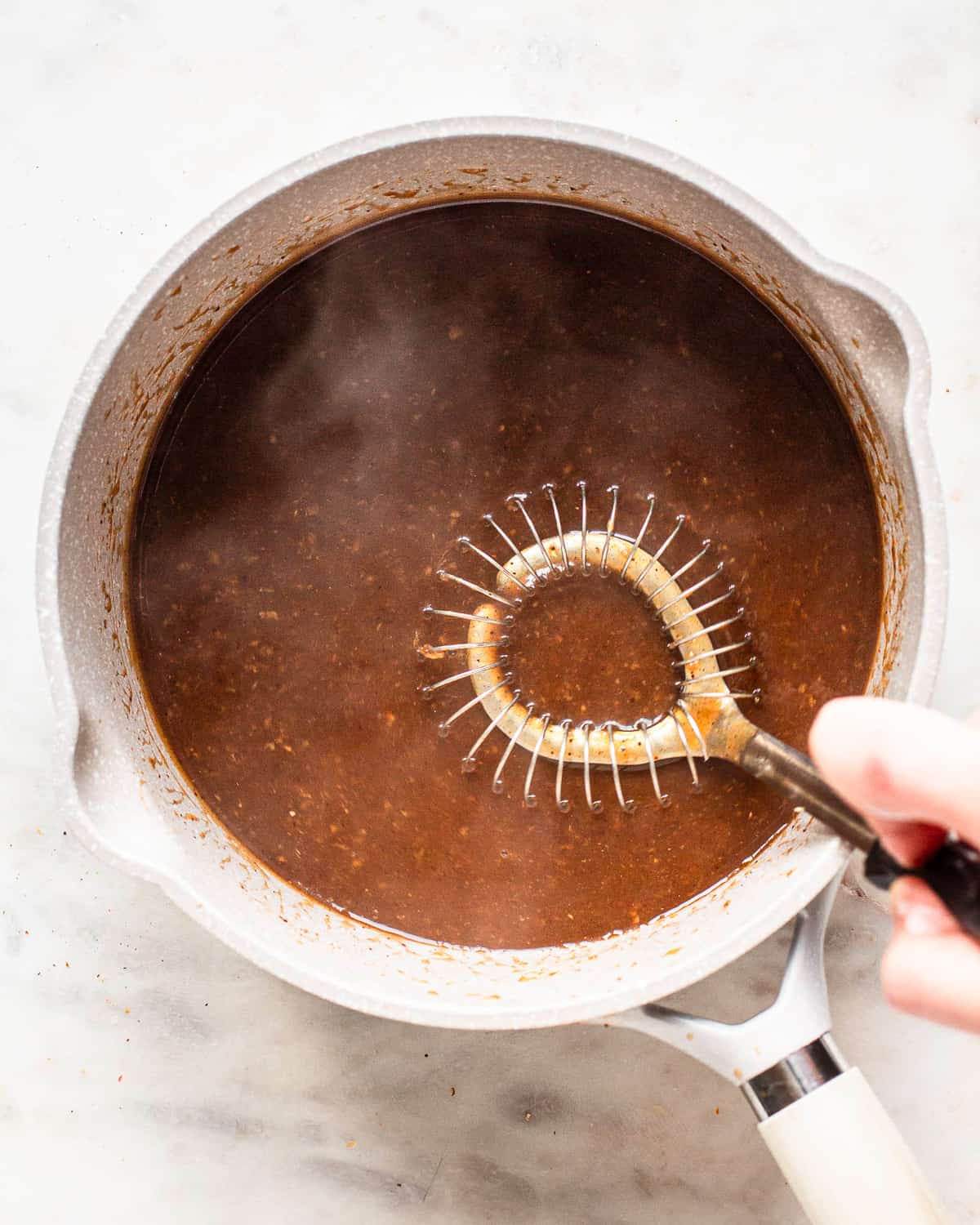 Hand whisking a brown sauce in a light grey saucepan.