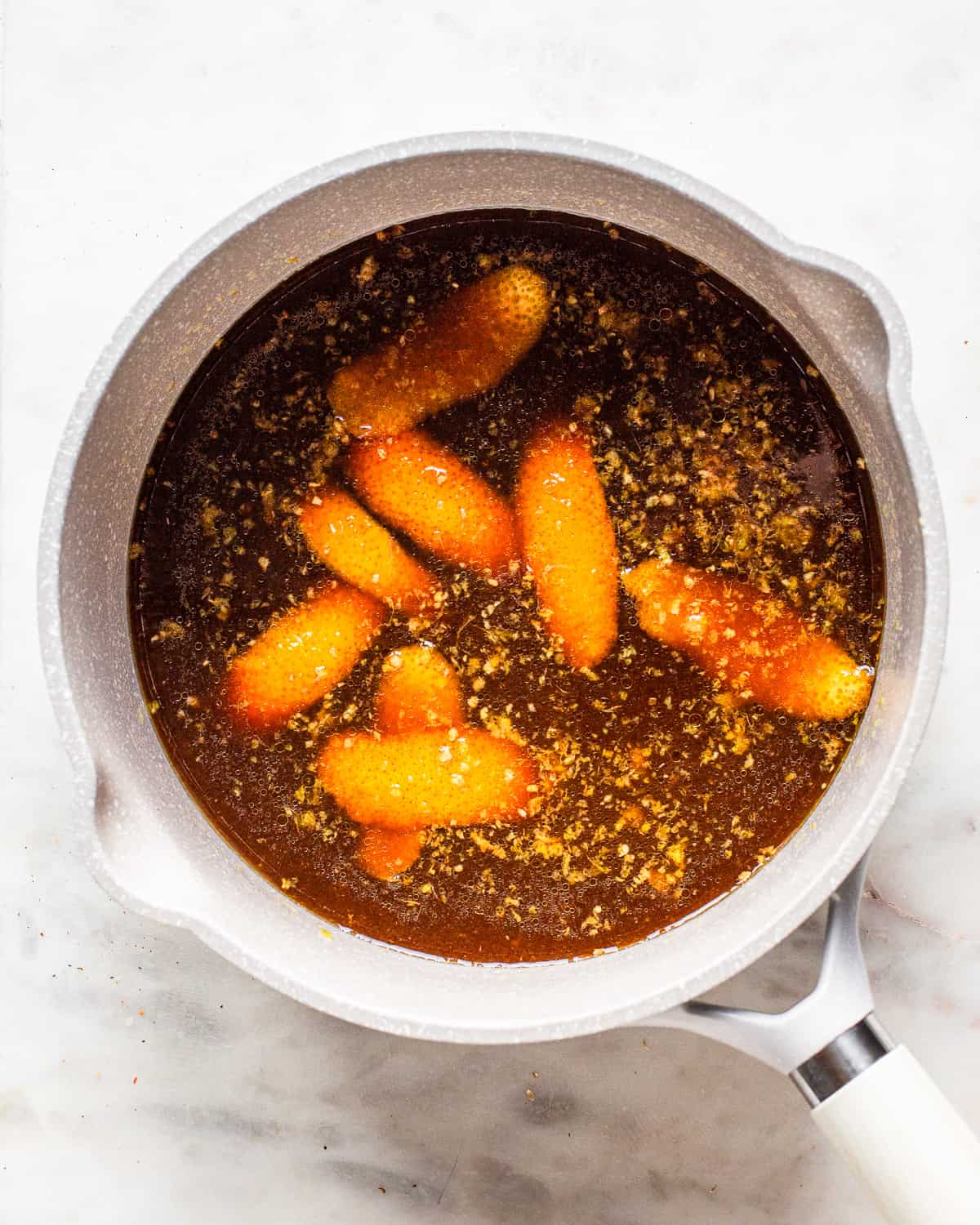 Light grey saucepan with orange zest swimming in a brown liquid.