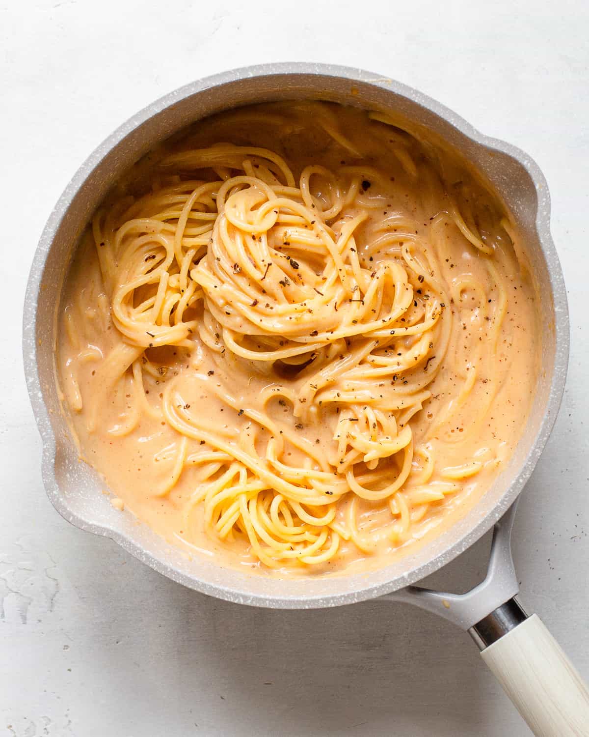 Cooked spaghetti tossed in a light orange sauce in a light grey saucepan.