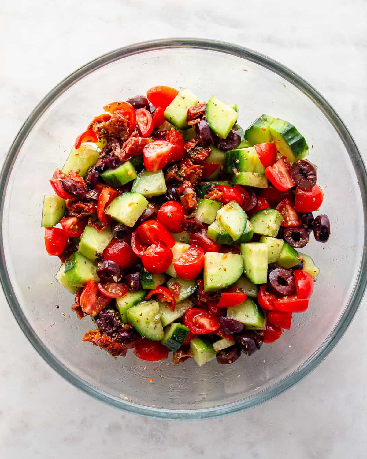 Glass bowl with seasoned cherry tomatoes, cucumber chunks, olives, and sun-dried tomatoes.