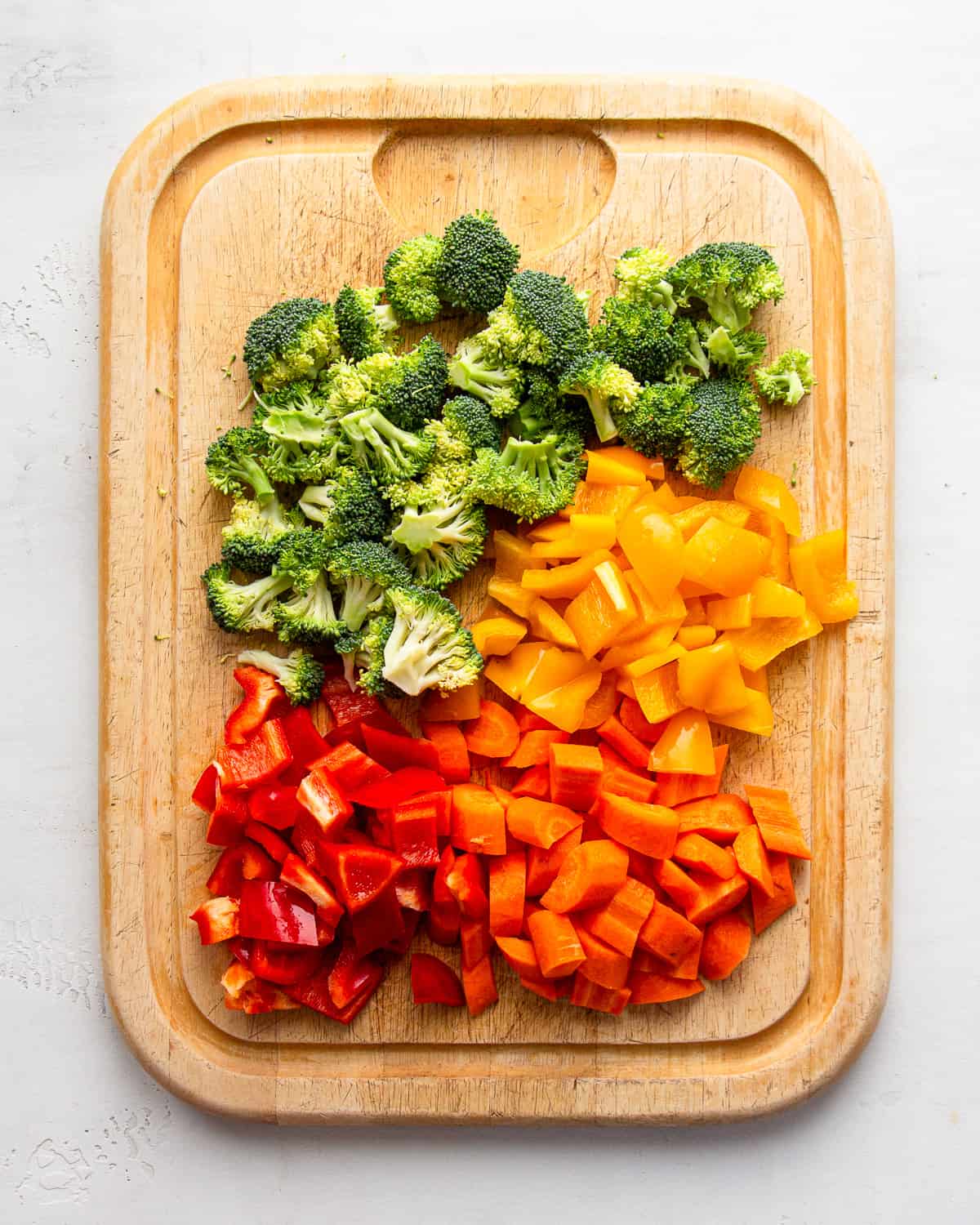 Chopped broccoli, carrots, red and orange bell peppers on a wooden cutting board.