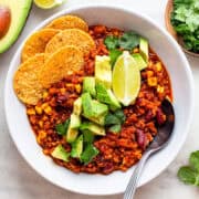 White bowl with vegan TVP chili, avocado, cilantro, and tortilla chips.