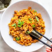 Hand pinching kimchi fried rice with a piece of nori with black chopsticks in a white bowl.